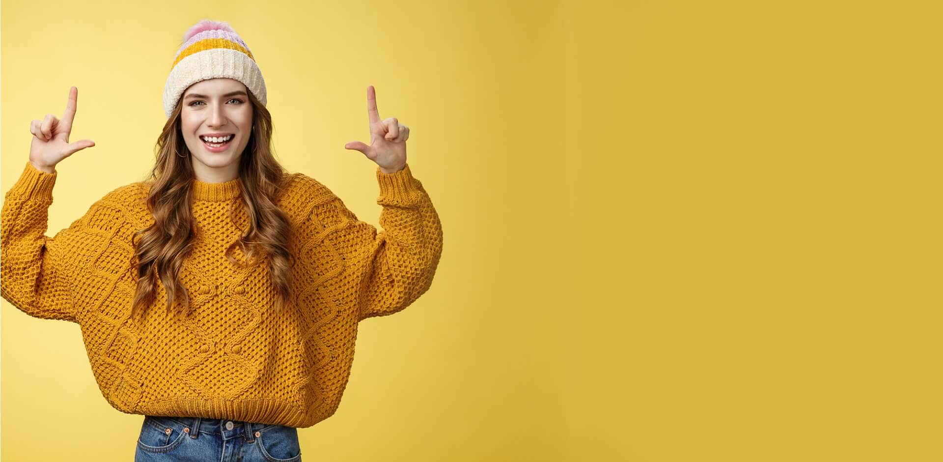A cheerful young woman in a yellow knit sweater and colorful beanie, smiling and pointing upwards, representing the energetic and customizable knitwear available from a custom sweater manufacturer