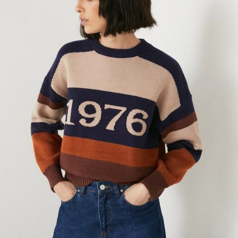 Woman wearing a custom 1976 wool-cashmere blend striped sweater in beige, navy, orange, and brown, paired with blue jeans.