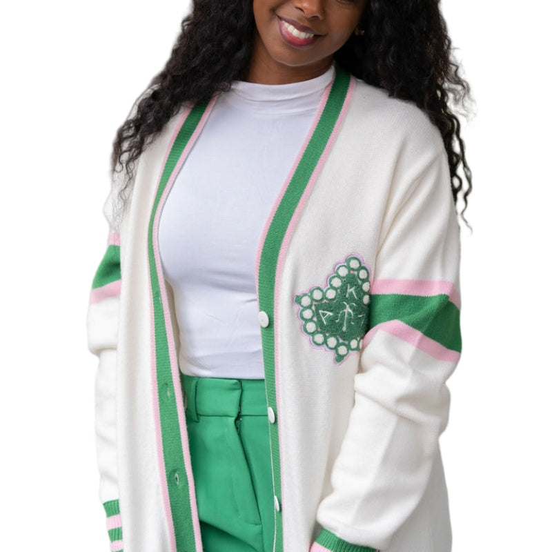 Confident African American woman smiling in a custom white Greek 100% cotton cardigan, detailed with green and pink stripes and an embroidered emblem on the chest. She is wearing a white t-shirt and bright green trousers, presenting a casual yet stylish appearance.