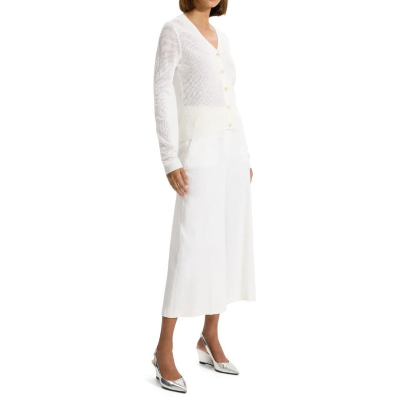 Full body view of a woman wearing a custom crochet acrylic knit sweater in white. The cardigan features a V-neckline, button-front design, and long sleeves, paired with white wide-leg pants and silver heels.