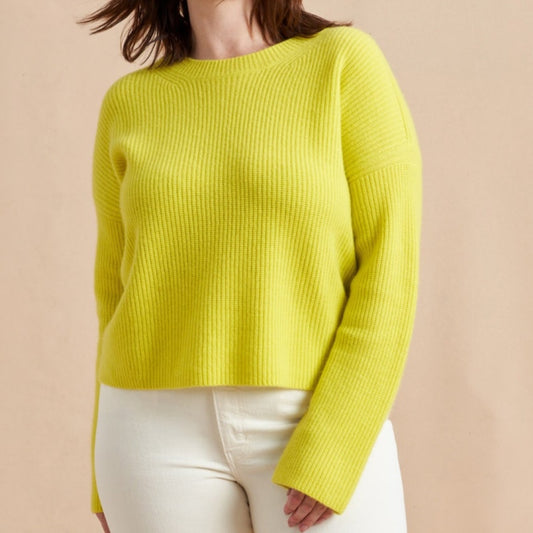 A woman wearing a bright yellow ribbed knit sweater with long sleeves and a round neckline, paired with a matching yellow skirt. The background is a neutral beige color.