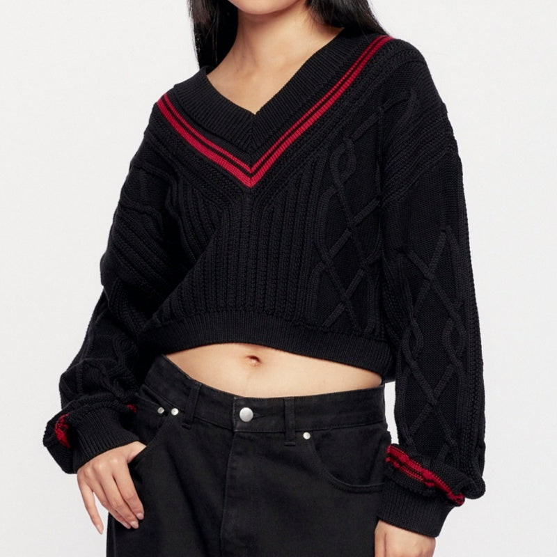 Front view of a woman wearing a black V-neck knitted sweater with red accents and intricate cable knit patterns, paired with black pants.