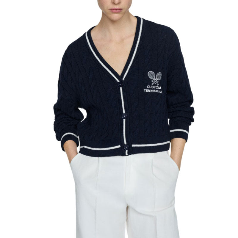 Person wearing a dark blue cable knit cardigan with custom tennis club logo embroidered on the chest, paired with white pants.