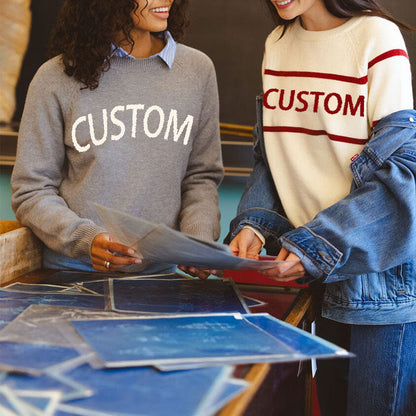 Two women discussing designs, one wearing a grey sweater with 'CUSTOM' in white and the other wearing a white sweater with red stripes and 'CUSTOM' in red.