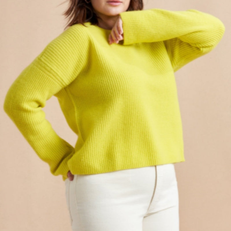 A woman wearing a bright yellow ribbed knit sweater with long sleeves and a round neckline, paired with a matching yellow skirt. The background is a neutral beige color.