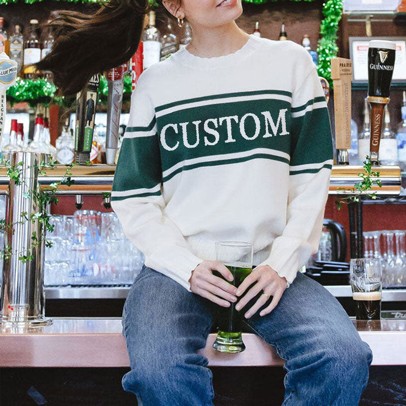 A woman sitting at a bar wearing a custom white knit sweater with green stripes and the word 'CUSTOM' holding a drink.