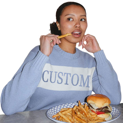 A woman wearing a light blue custom knit sweater with a white stripe and the word 'CUSTOM' eating fries and a burger.