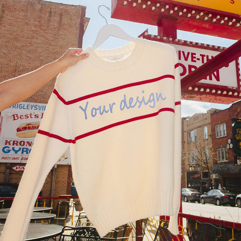 Hand holding a white knit sweater with red horizontal stripes and the text 'Your design' written in blue across the chest, with a city street background.