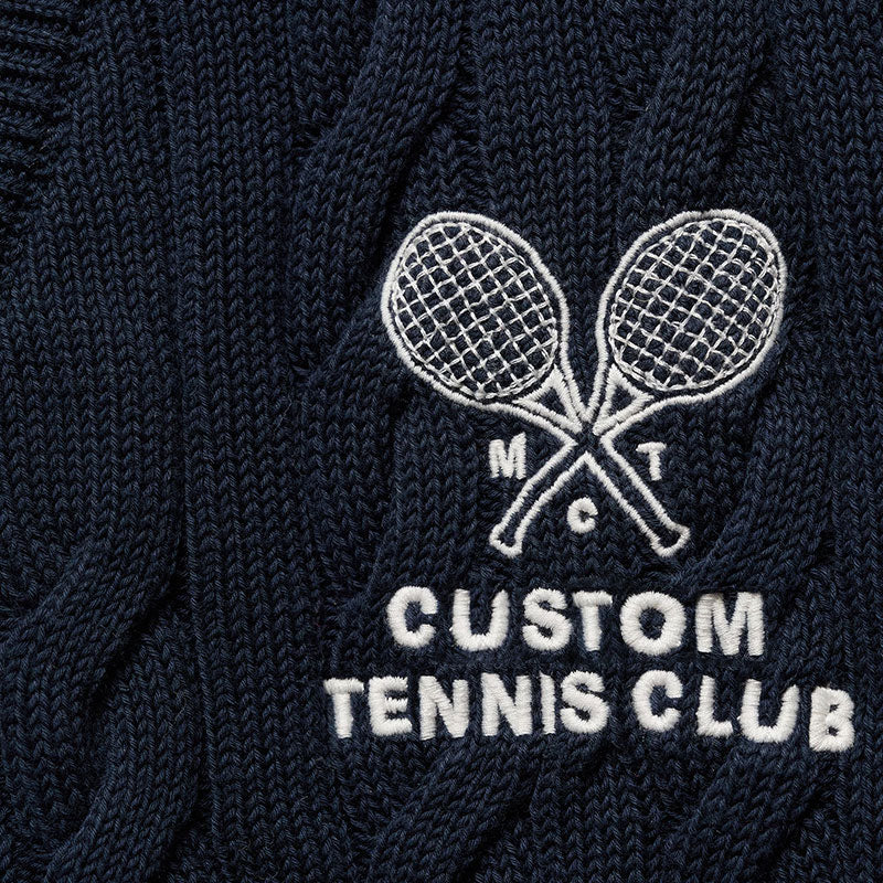 Close-up of the custom tennis club logo embroidered on a dark blue cable knit cardigan, featuring two crossed tennis rackets.