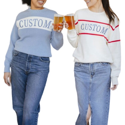 Two women wearing custom knit sweaters, one in light blue with a white stripe and the word 'CUSTOM' and the other in white with red stripes and the word 'CUSTOM', holding beers.