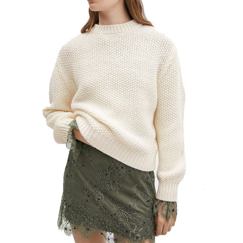 Model wearing a cream-colored, grid pullover sweater paired with a green lace skirt. The sweater has a textured knit pattern, and the model has one sleeve rolled up.