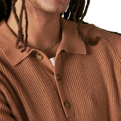 Close-up of the camel-colored ribbed knit polo sweater, focusing on the button placket and collar detail, worn by a man with dreadlocks.