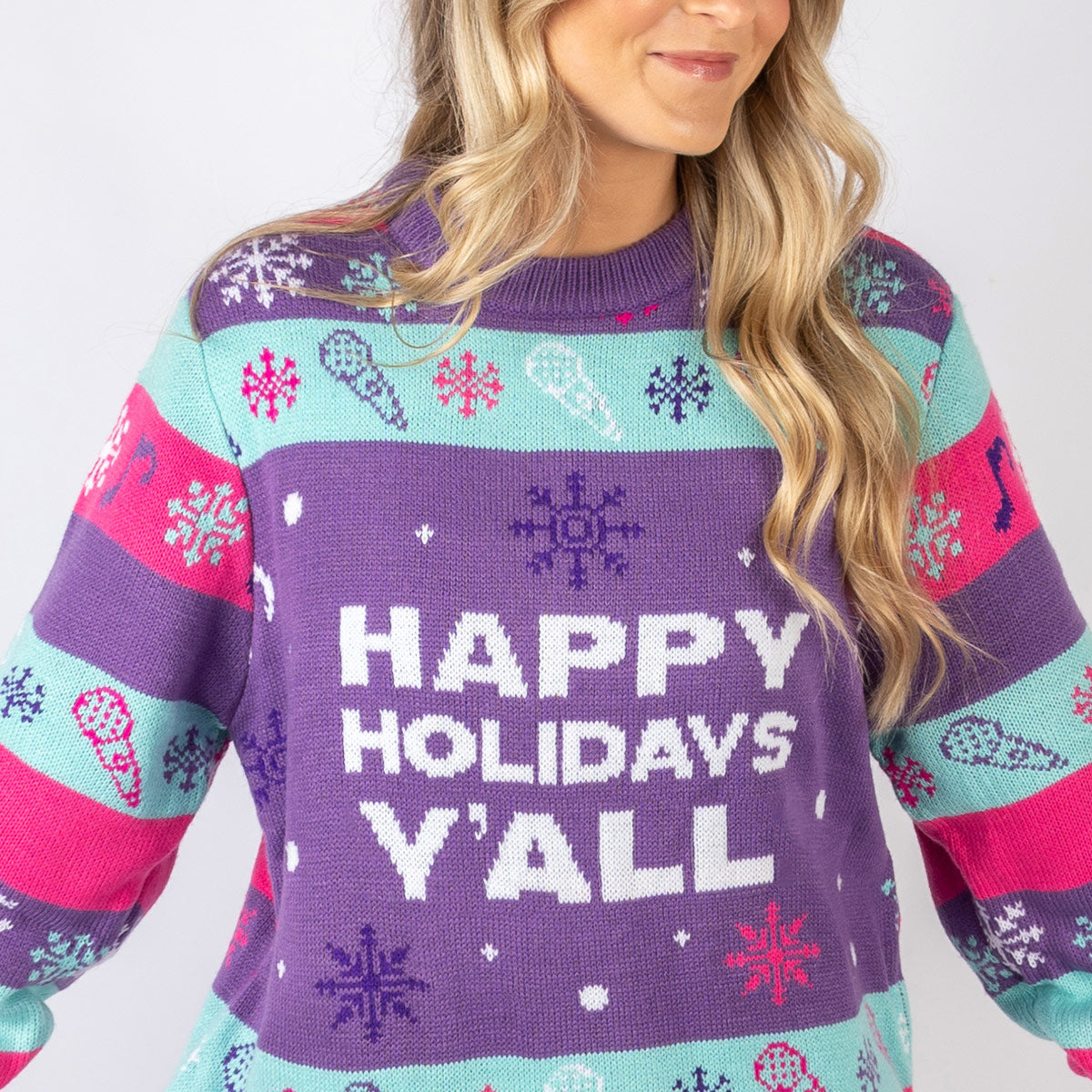 Model wearing a custom cozy cashmere Christmas sweater with a 'Happy Holidays Y'all' design in purple, pink, teal, and white colors. The sweater features holiday motifs including snowflakes, musical notes, and festive decorations.