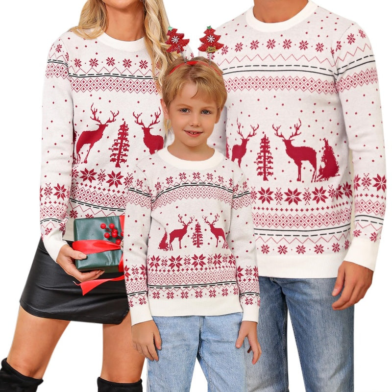 A happy family of three wearing matching custom knit wool Christmas sweaters with red reindeer and snowflake patterns. The mother is holding a gift, and the child is standing in front with a smile.