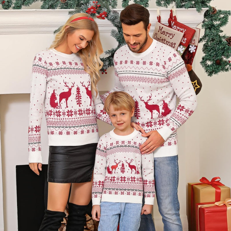 A family of three celebrating Christmas in front of a decorated fireplace, all wearing matching custom knit wool sweaters with red reindeer and snowflake patterns. The parents are looking at their child who stands in front.