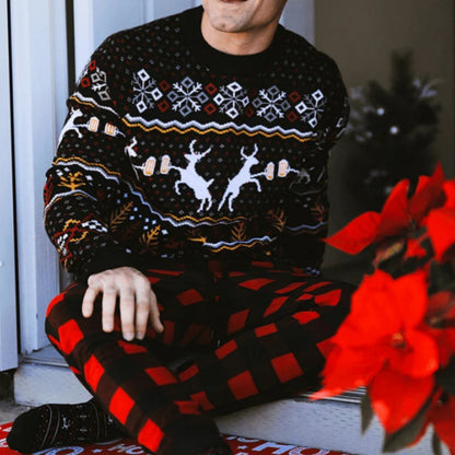 A man sitting on the floor in a cozy setting, wearing a festive ugly Christmas sweater with reindeer and geometric patterns, paired with red and black checkered pants. A Christmas tree and red poinsettia flowers are in the background.